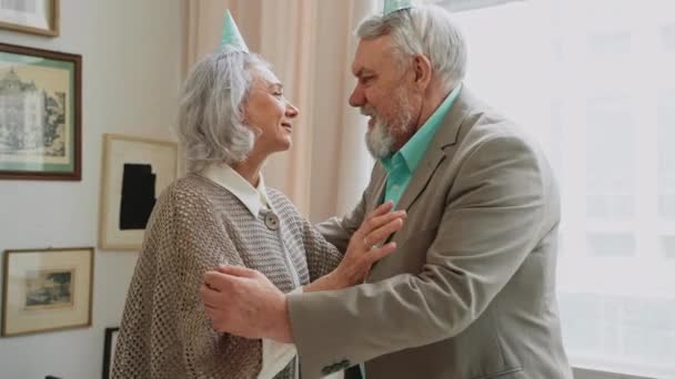 Vanhukset halailevat ja tervehtivät toisiaan syntymäpäiväjuhlissa. Pastellivärisiä vaatteita senioreille. Harmaahiuksinen nainen halaamassa miestään. Laadukas 4k kuvamateriaalia - Materiaali, video