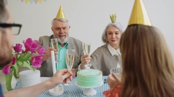 Gelukkige oudere paar clink glazen met familie op een helder verjaardagsfeest. Pastel gekleurde partij met kegels op hoofden. Ouderen op een feestje. Hoge kwaliteit 4k beeldmateriaal - Video