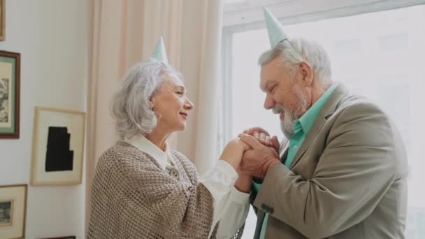 Ouderen knuffelen en zoenen elkaar op het verjaardagsfeest. Een man met grijs haar kust zijn vrouw haar handen. Pastelkleurige kleding op senioren. Hoge kwaliteit 4k beeldmateriaal - Video