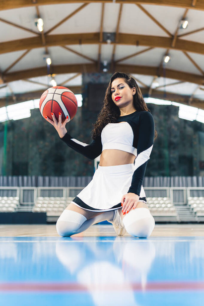 Cheerleader donna seduta sulle ginocchia e tenendo una pallacanestro in su in una mano sul campo. Foto di alta qualità - Foto, immagini