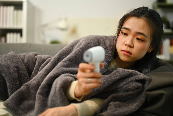 Mujer joven en manta acostada en el sofá y midiendo la fiebre con termómetro infrarrojo. Concepto sanitario. - Foto, Imagen