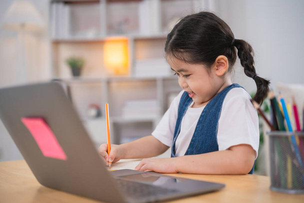Ázsiai kislány mosolyog használja laptop vagy írás rajz színe jegyzetfüzet tanulmány online fa asztal asztal nappaliban otthon. Oktatás online tanulás otthonról. - Fotó, kép