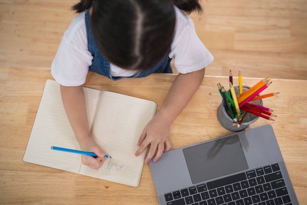 Топвью. Азиатская девочка улыбается использовать ноутбук или писать рисунок цвета на записной книжке исследование онлайн на деревянный стол стол в гостиной дома. Образование онлайн из домашней концепции. - Фото, изображение