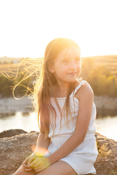 Милая маленькая девочка с длинными волосами на фоне реки в природе, улыбается, смотрит в сторону. - Фото, изображение
