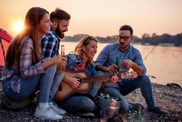 Groep vrienden zitten en luisteren naar de muziek tijdens het drinken van cider bij zonsondergang. - Foto, afbeelding