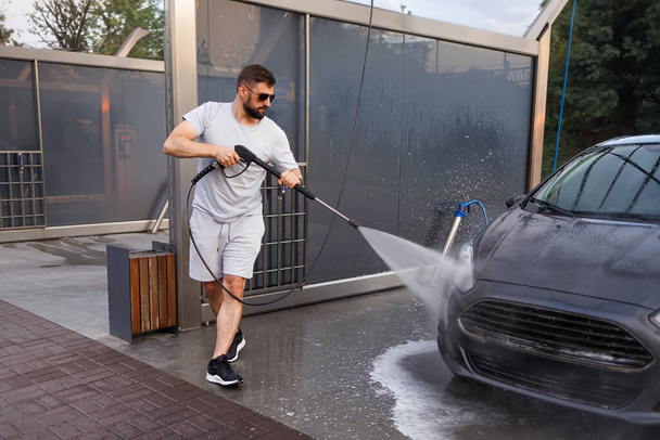 Bir adam bir arabanın ön ızgarasını su topuyla yıkıyor. Otomobil yıkamada bir araba.. - Fotoğraf, Görsel