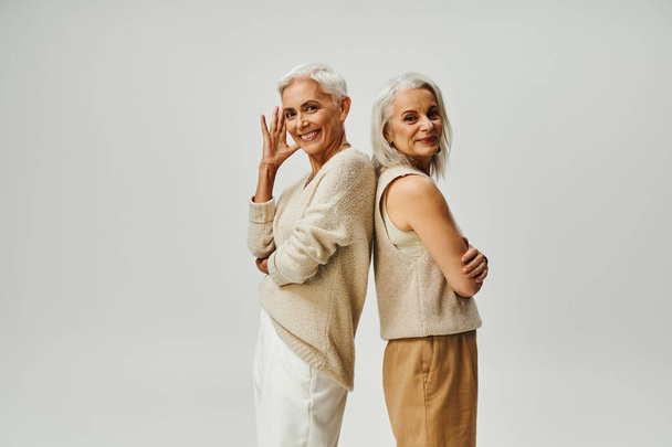 aging gracefully, stylish senior women standing back to back and smiling at camera on grey - Photo, Image