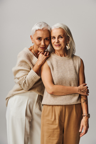 mujer madura apoyada en el hombro de una amiga feliz mirando a la cámara en gris, envejecimiento agraciado - Foto, imagen