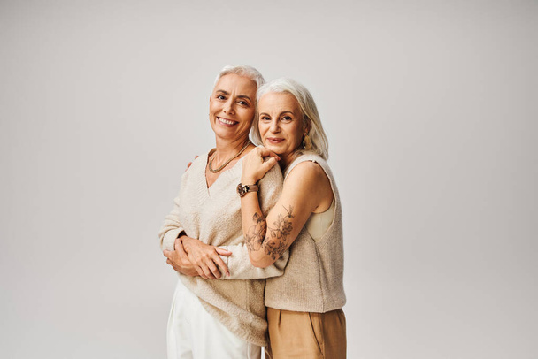 mature tattooed lady hugging cheerful and fashionable female friend on grey, positive aging - Photo, Image