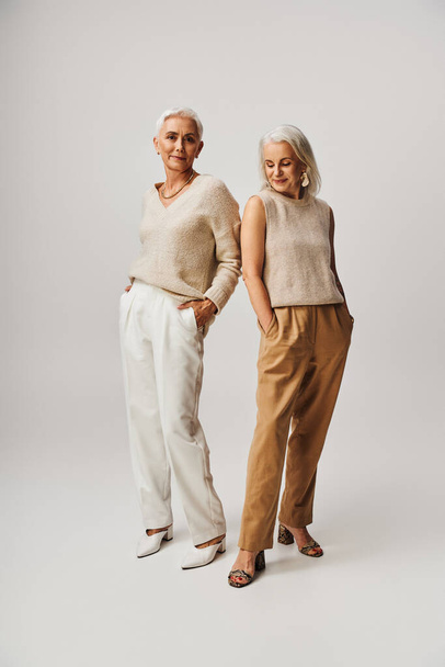 positieve volwassen dames in stijlvolle casual kleding rusten met de handen in zakken op grijs, volledige lengte - Foto, afbeelding