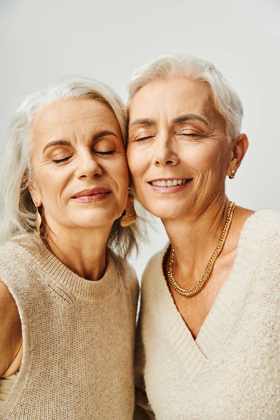 χαρούμενες ηλικιωμένες γυναίκες με χρυσά αξεσουάρ και μακιγιάζ με κλειστά μάτια σε γκρι, δια βίου φίλους - Φωτογραφία, εικόνα