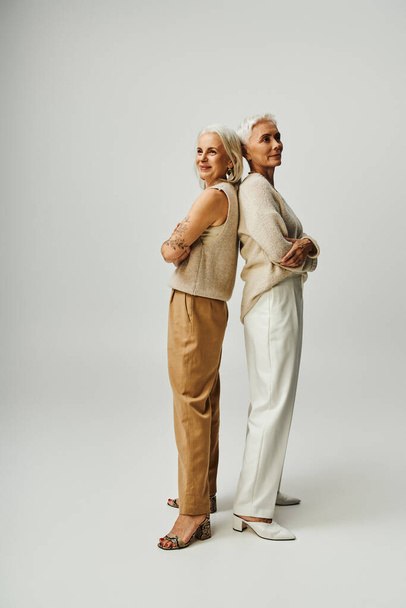 full length of senior fashionistas standing back to back with folded arms on grey, lifestyle friends - Photo, Image
