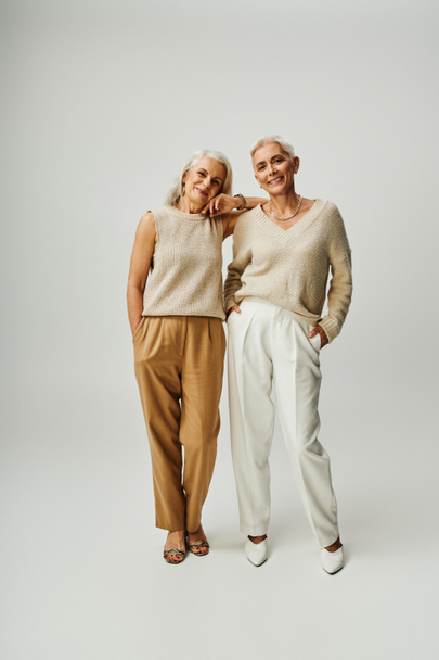 volle Länge der Smiley-Senior-Models in pastellfarbener Freizeitkleidung stehend mit Händen in Taschen auf grau - Foto, Bild