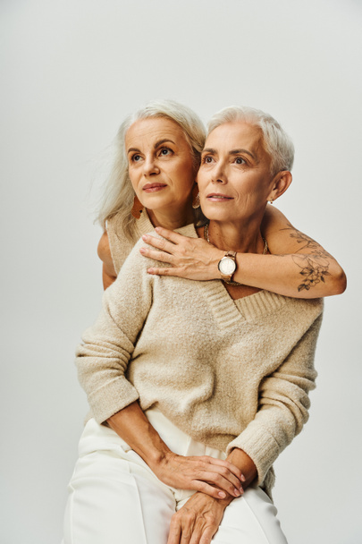 starší tetovaná žena objímající zasněnou přítelkyni dívající se jinam na šedý, elegantní životní styl - Fotografie, Obrázek
