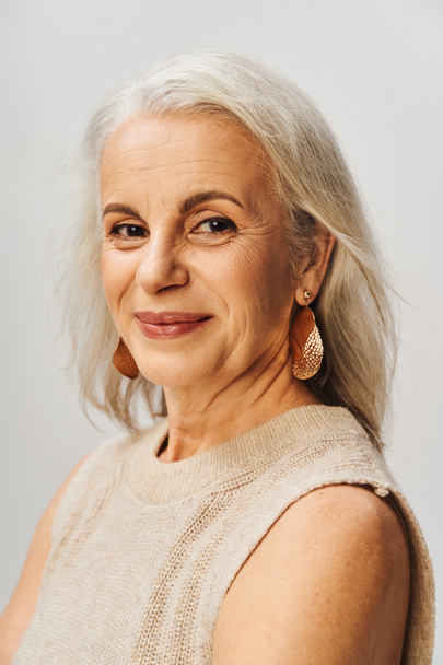 portrait de femme âgée heureuse en maquillage et boucles d'oreilles dorées regardant la caméra sur fond gris - Photo, image