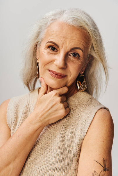 portrét stříbrovlasého staršího modelky v make-upu a zlatých náušnicích s úsměvem na kameru na šedi - Fotografie, Obrázek