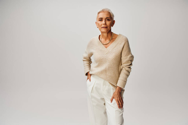 zelfverzekerde senior fashionista met zilver haar poseren met de hand in zak van witte broek op grijs - Foto, afbeelding