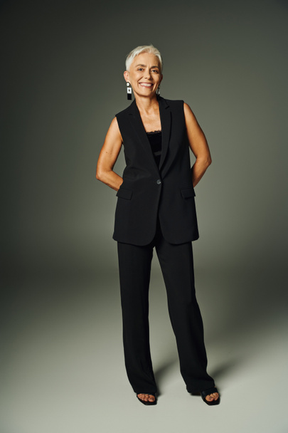 mulher sênior na moda com sorriso radiante posando em traje elegante preto em cinza, comprimento total - Foto, Imagem