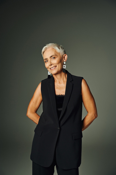 smiley senior woman with short silver hair posing in black attire with hands behind back on grey - Photo, Image