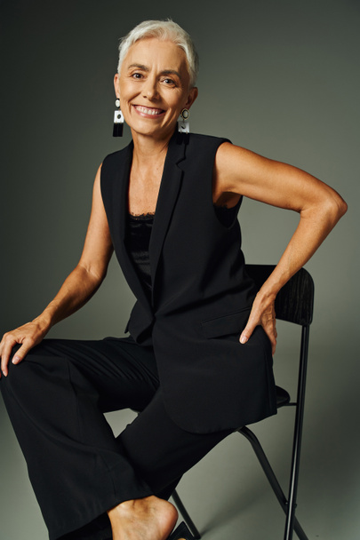 stárnutí elegantně, usmívající se starší žena v černém oblečení sedí na židli s rukou na boku na šedé - Fotografie, Obrázek