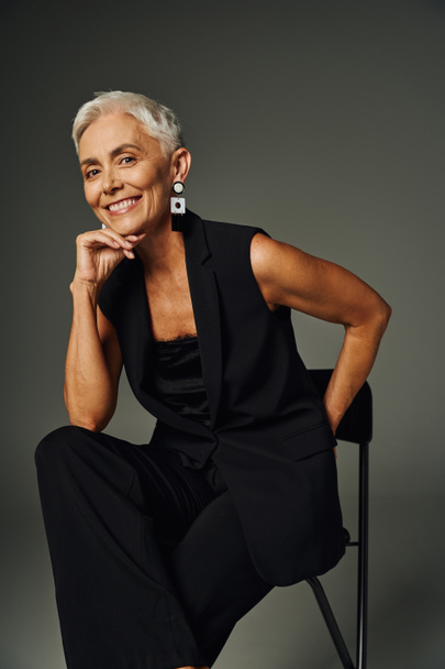 θετική, χαρούμενη ηλικιωμένη γυναίκα με μαύρη ενδυμασία κάθεται στην καρέκλα με το χέρι πίσω από την πλάτη σε γκρι - Φωτογραφία, εικόνα
