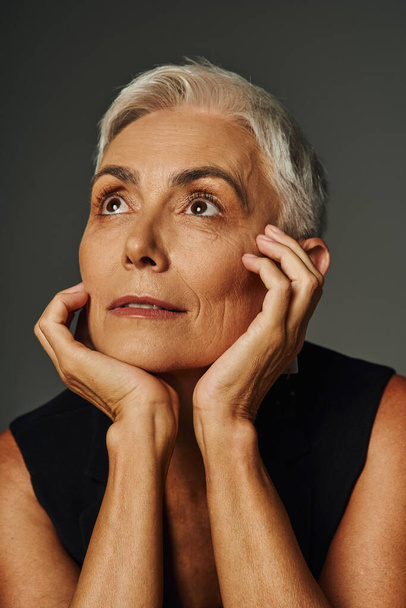 портрет мечтательной пожилой женщины с короткими серебряными волосами, смотрящей в сторону с руками рядом с лицом на сером - Фото, изображение
