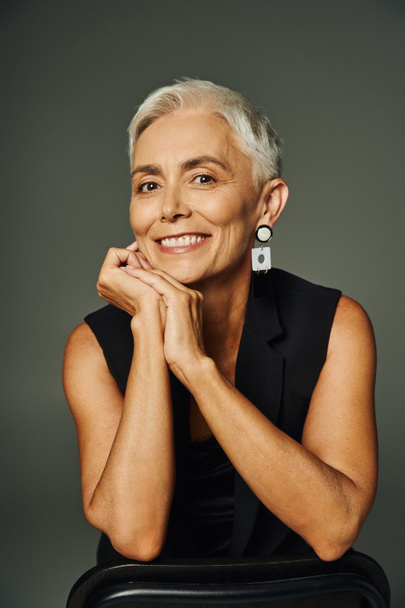 portrait of trendy senior lady posing with hands near face and smiling at camera on grey backdrop - Photo, Image