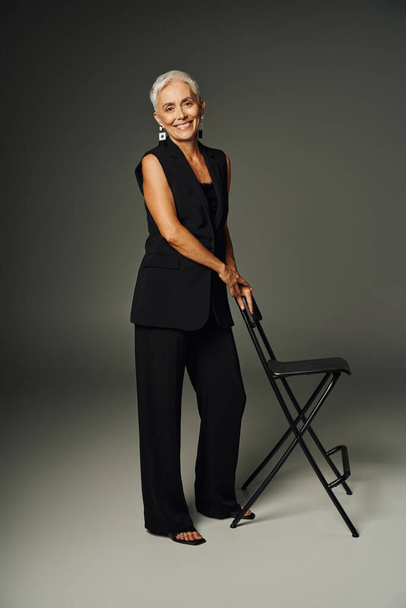 plná délka radostné starší dámy v černém klasickém oblečení pózování s židlí na šedé, elegance - Fotografie, Obrázek