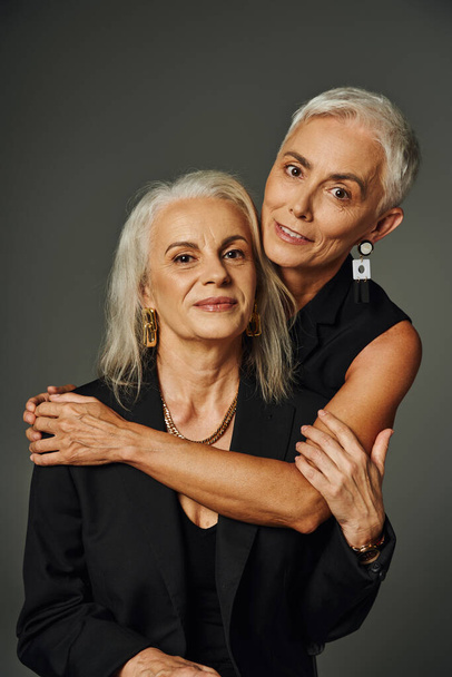 fashionable senior lady in black attire embracing trendy female friend on grey, elegant aging - Photo, Image