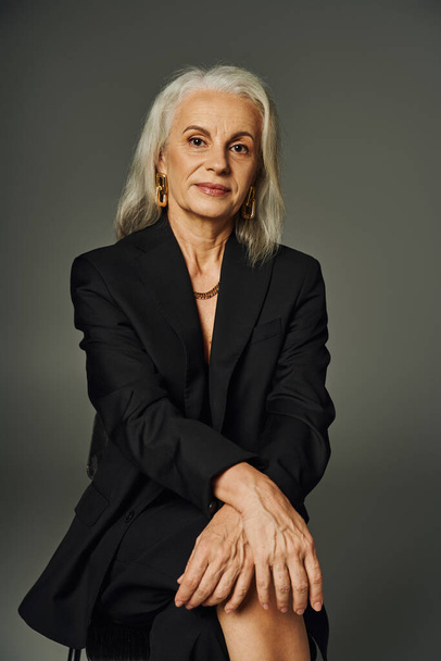 ältere weibliche Modell in schwarzer eleganter Kleidung sitzt und blickt ta Kamera auf graue, anmutige Alterung - Foto, Bild