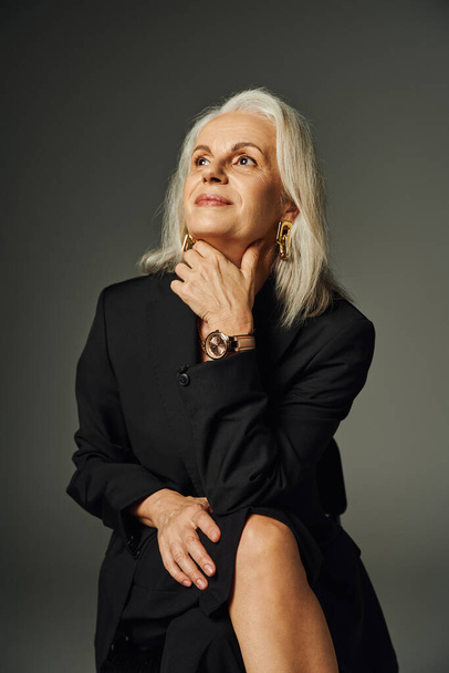 femme mature rêveuse et souriante en noir élégant porter assis et regardant loin sur gris, élégance - Photo, image