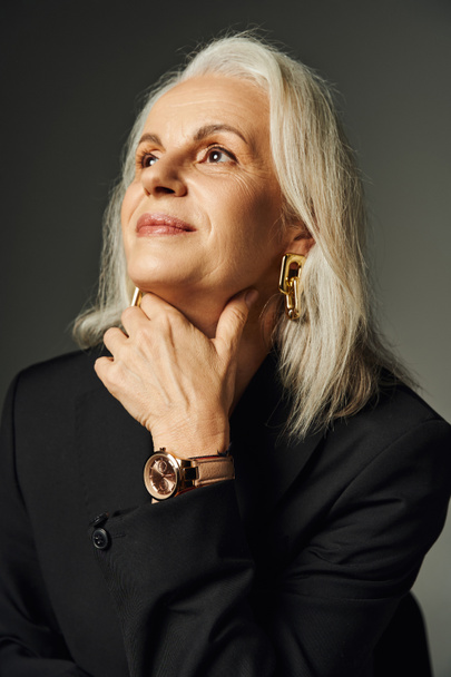 портрет мечтательной и элегантной пожилой женщины в наручных часах и золотых аксессуарах, смотрящей в сторону на серый - Фото, изображение