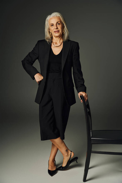 longitud completa del modelo senior en traje negro posando con la mano en la cadera cerca de la silla en gris, elegancia - Foto, Imagen