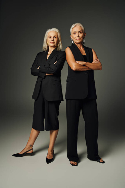 ανώτερος φωτογραφία μόδας, κομψές κυρίες σε μαύρα ρούχα ποζάρουν με διπλωμένα χέρια σε γκρι φόντο - Φωτογραφία, εικόνα