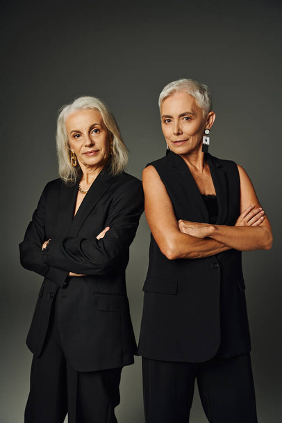 trendy senior models posing with folded arms in black classis attire and looking at camera on black - Photo, Image