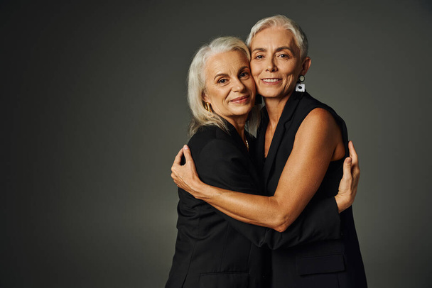 amigos de toda la vida, señoras mayores alegres y elegantes en ropa negra abrazando en fondo gris - Foto, Imagen