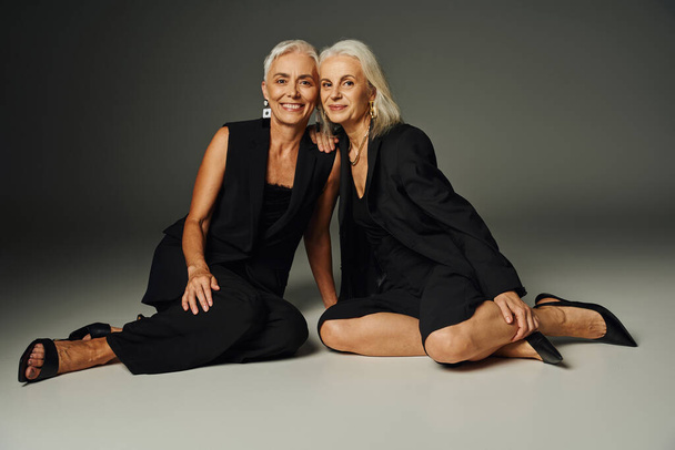 modelos seniores encantadores e felizes em traje clássico preto sentado em pano de fundo cinza, estilo justo vaidade - Foto, Imagem