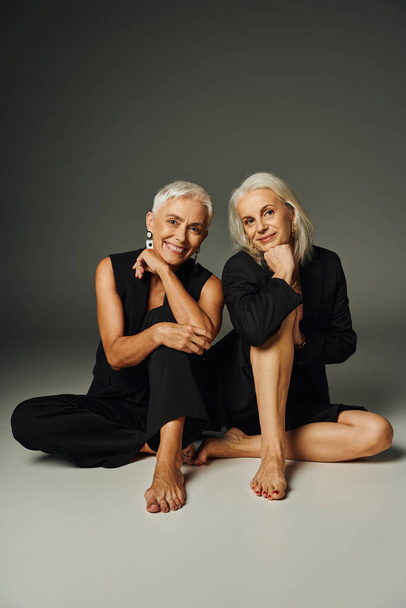 cheerful barefoot senior models in black fashionable clothes sitting and looking at camera on grey - Photo, Image