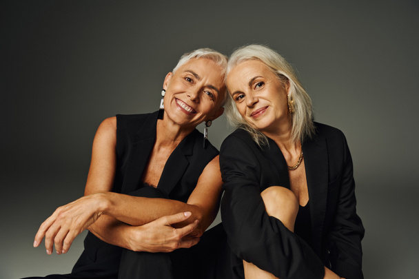 amies aînées en tenue noire à la mode souriant à la caméra tout en posant sur gris, positivité - Photo, image