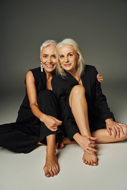 happy and trendy barefoot senior women in black clothes looking at camera while sitting on grey - Photo, Image
