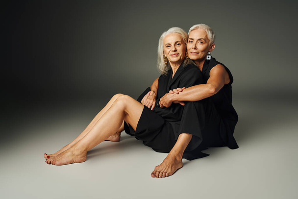 happy and barefoot senior women in black attire sitting and smiling at camera on grey, age positive - Photo, Image