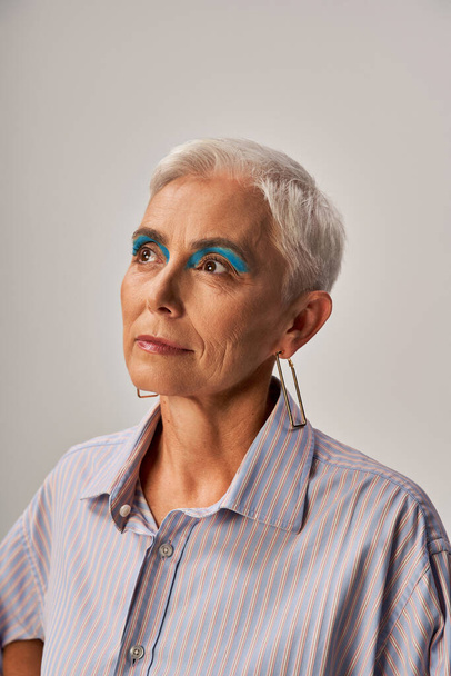 stijlvol dromerig senior model met kort zilver haar en blauwe eyeliner weg te kijken op grijs, portret - Foto, afbeelding