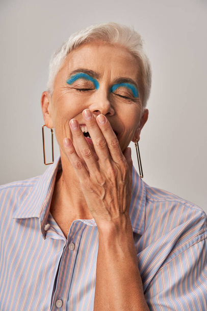 возбужденная зрелая женщина с смелым макияжем смеется с закрытыми глазами и закрывая рот рукой на сером - Фото, изображение