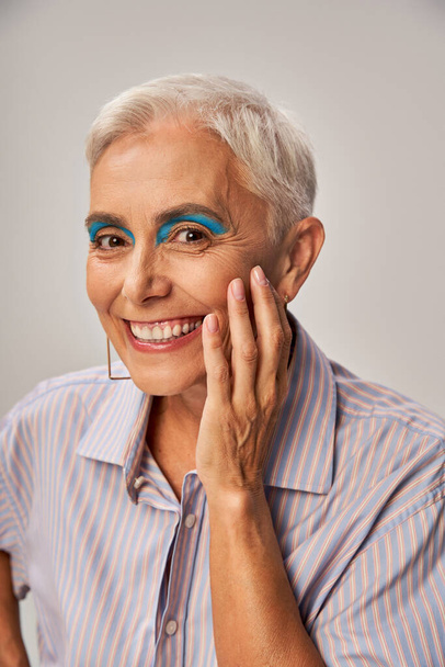 портрет стильной старшей модели с короткими серебряными волосами и смелым макияжем, улыбающимся в камеру на сером - Фото, изображение