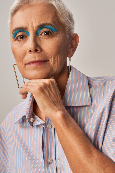 dame mature à la mode avec les cheveux courts argentés et eye-liner bleu posant avec la main près du menton sur gris - Photo, image