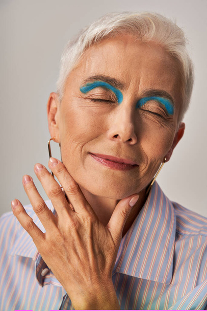 mujer madura positiva con pelo corto plateado y delineador azul posando con los ojos cerrados sobre gris - Foto, Imagen