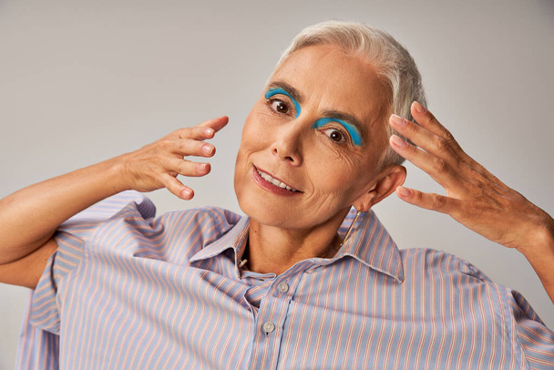 modelo de moda senior con delineador de ojos azul y pelo corto plateado con las manos cerca de la cara en gris, retrato - Foto, Imagen