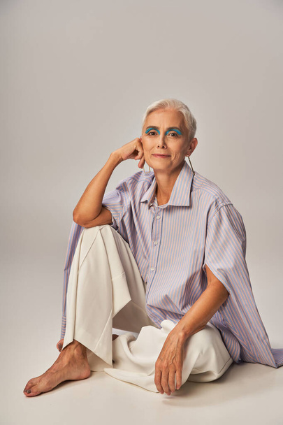 модная босиком старшая женщина в голубой полосатой рубашке и белых брюках, сидящих на сером фоне - Фото, изображение