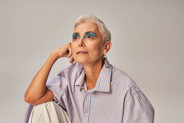 pensive en stijlvolle senior vrouw in blauw gestreept shirt en gedurfde make-up weg te kijken op grijs - Foto, afbeelding