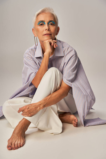 trendige alternde, barfüßige reife Frau im blau gestreiften Hemd sitzend und in die Kamera blickend auf grau - Foto, Bild